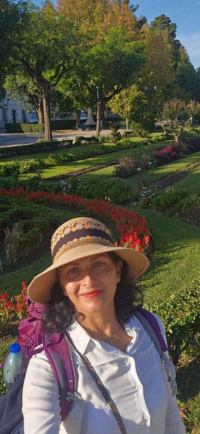 Mit Lida Pilgern - Pilgerreise-Coaching - Eine Frau mit Sonnenhut und Rucksack macht ein Selfie in einem üppigen, terrassenförmig angelegten Garten mit leuchtenden Blumen und grünen Bäumen unter klarem, blauem Himmel.