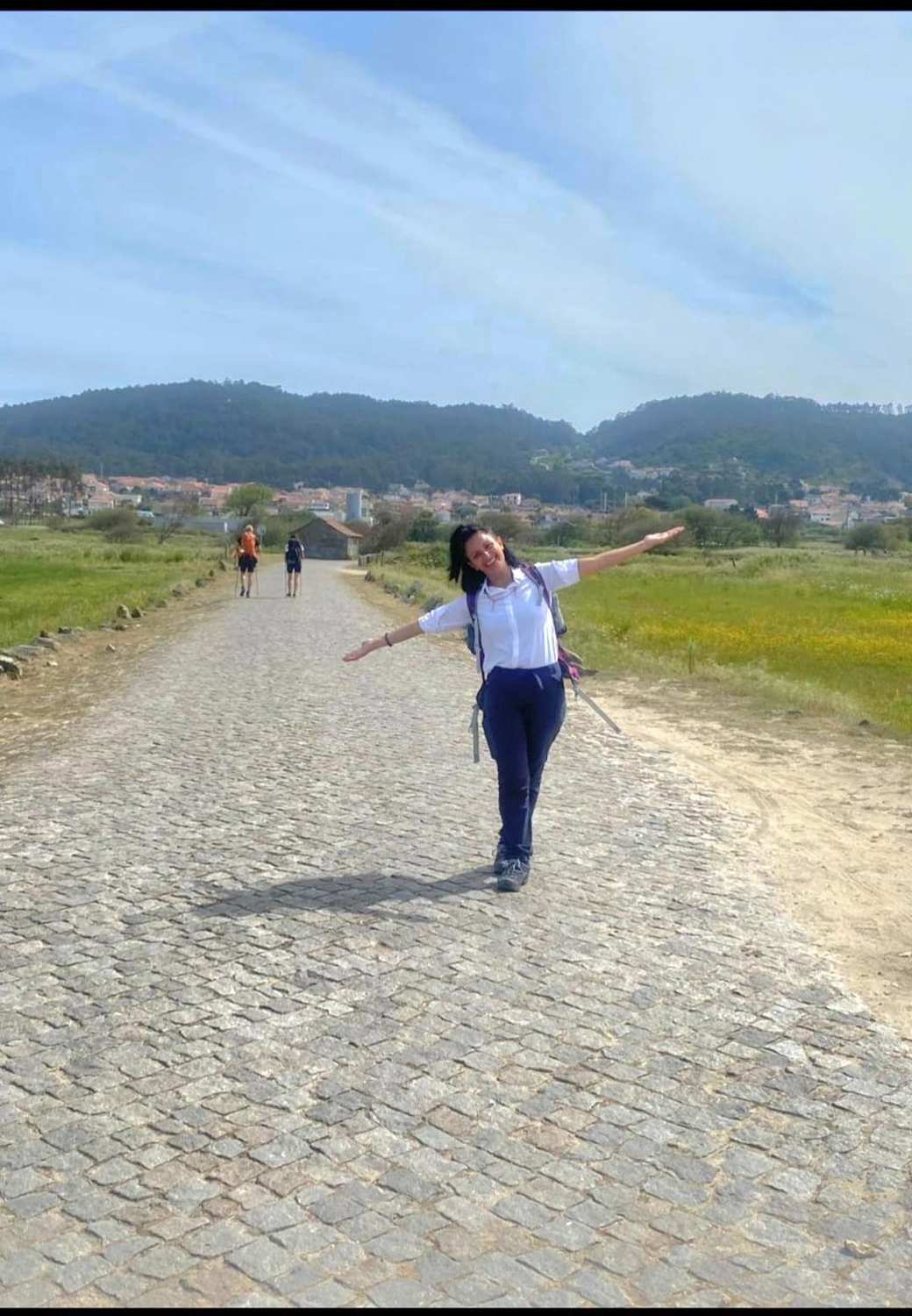 Mit Lida Pilgern - Pilgerreise-Coaching - Eine Person steht mit ausgestreckten Armen auf einer langen Kopfsteinpflasterstraße, die sich durch eine ländliche Landschaft unter einem hellen, leicht bewölkten Himmel schlängelt. Im Hintergrund sind grüne Felder, sanfte Hügel und zwei weitere Spaziergänger zu sehen.