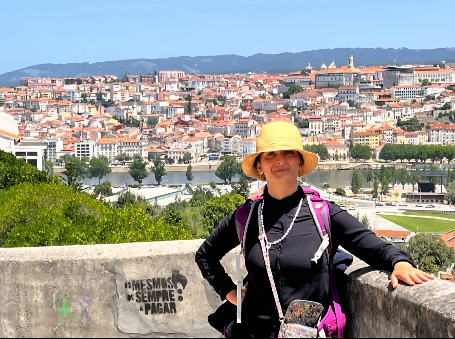 Mit Lida Pilgern - Pilgerreise-Coaching - Eine Frau mit gelbem Sonnenhut und schwarzem Hemd steht vor einer weitläufigen Hügelstadt mit rotgedeckten Gebäuden und einem Fluss.