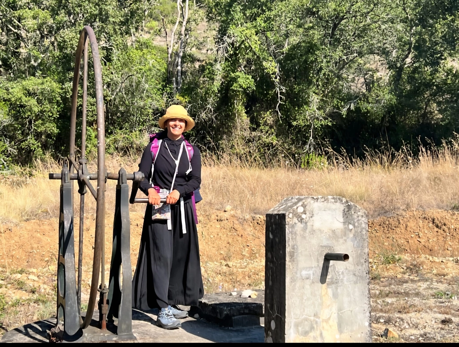 Mit Lida Pilgern - Pilgerreise-Coaching - Eine Frau in Wanderkleidung und Sonnenhut lächelt, während sie neben einem großen, rustikalen Eisenrad und einem Betonbauwerk in einer sonnigen, bewaldeten Umgebung steht.