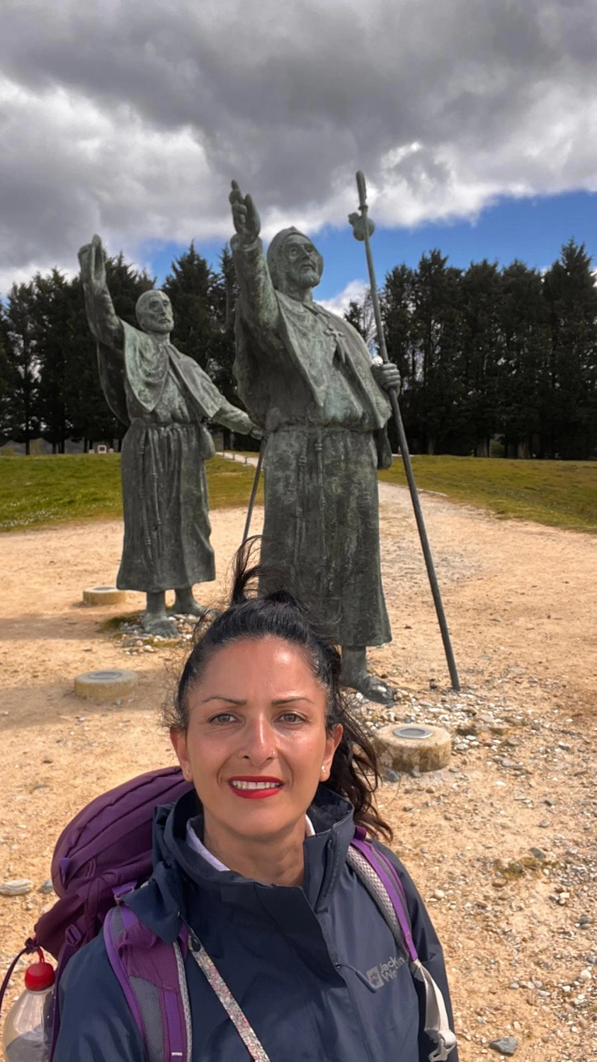 Mit Lida Pilgern - Pilgerreise-Coaching - Eine Frau in Wanderkleidung und Sonnenhut lächelt, während sie neben einem großen, rustikalen Eisenrad und einem Betonbauwerk in einer sonnigen, bewaldeten Umgebung steht.